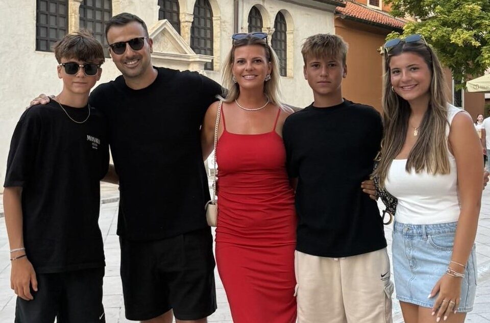 A group of five people, two adults and three teenagers, stand close together outdoors, smiling at the camera. The woman in the center wears a red dress; the others wear casual summer clothes in black and white.